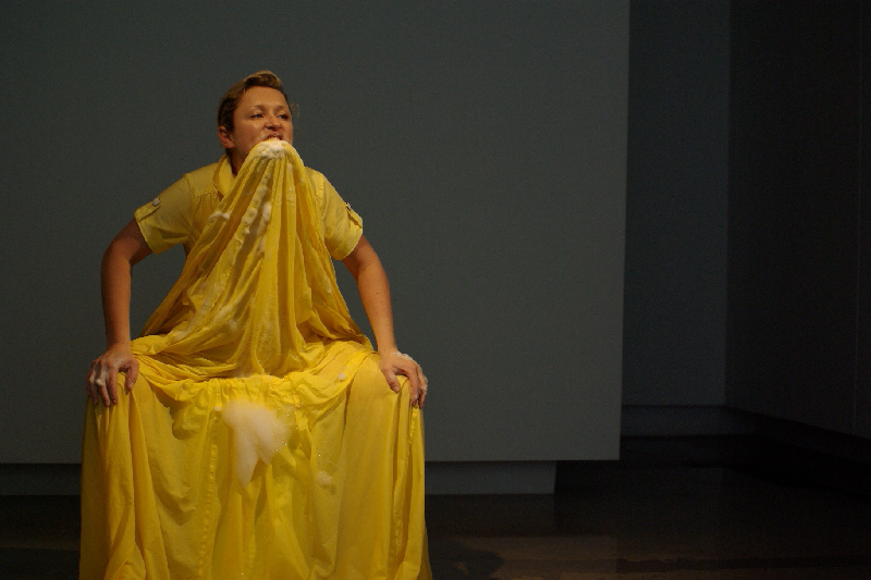 A woman is seated wearing a long, yellow dress with a full skirt. She has the front of the skirt held in her teeth, with her hands on her knees. There are some soap suds on her hands, near her mouth, and on the front of the skirt.