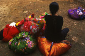 A person with a long braid is sitting on one of many brightly colored bundles of objects. The bundles are sitting on the dirt ground.