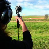 An over-the-shoulder photo of a person holding a boom microphone pointed at a small oil pipeline fire several dozen feet away.