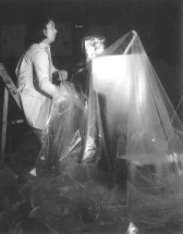 Black-and-white photograph of a figure, in profile, sitting behind a reed organ that is covered in plastic.