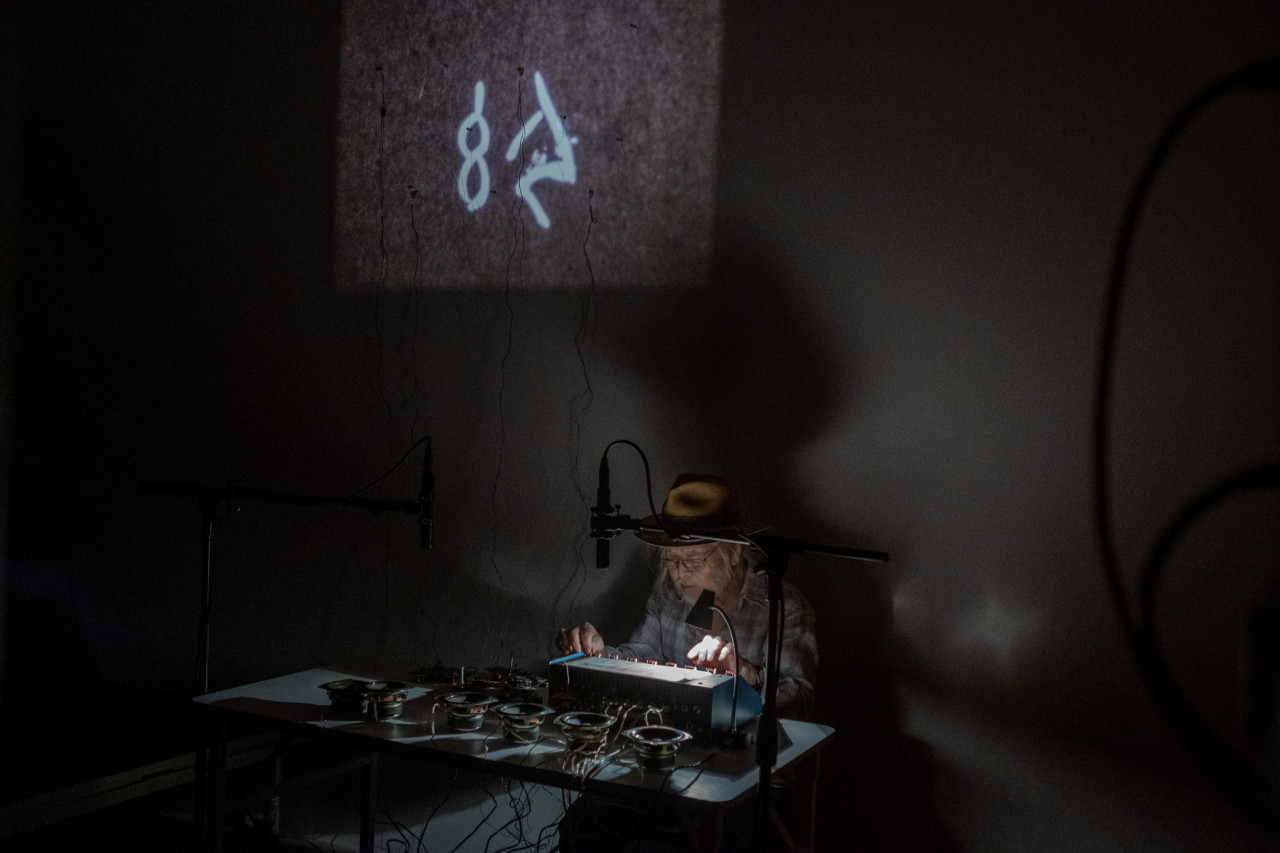 Color image of a darkly lit room. A figure sits behind a white table filled with audio equipment and two standing microphones are placed above the table. Light sensors are attached to the wall behind the figure where a projection of images plays.