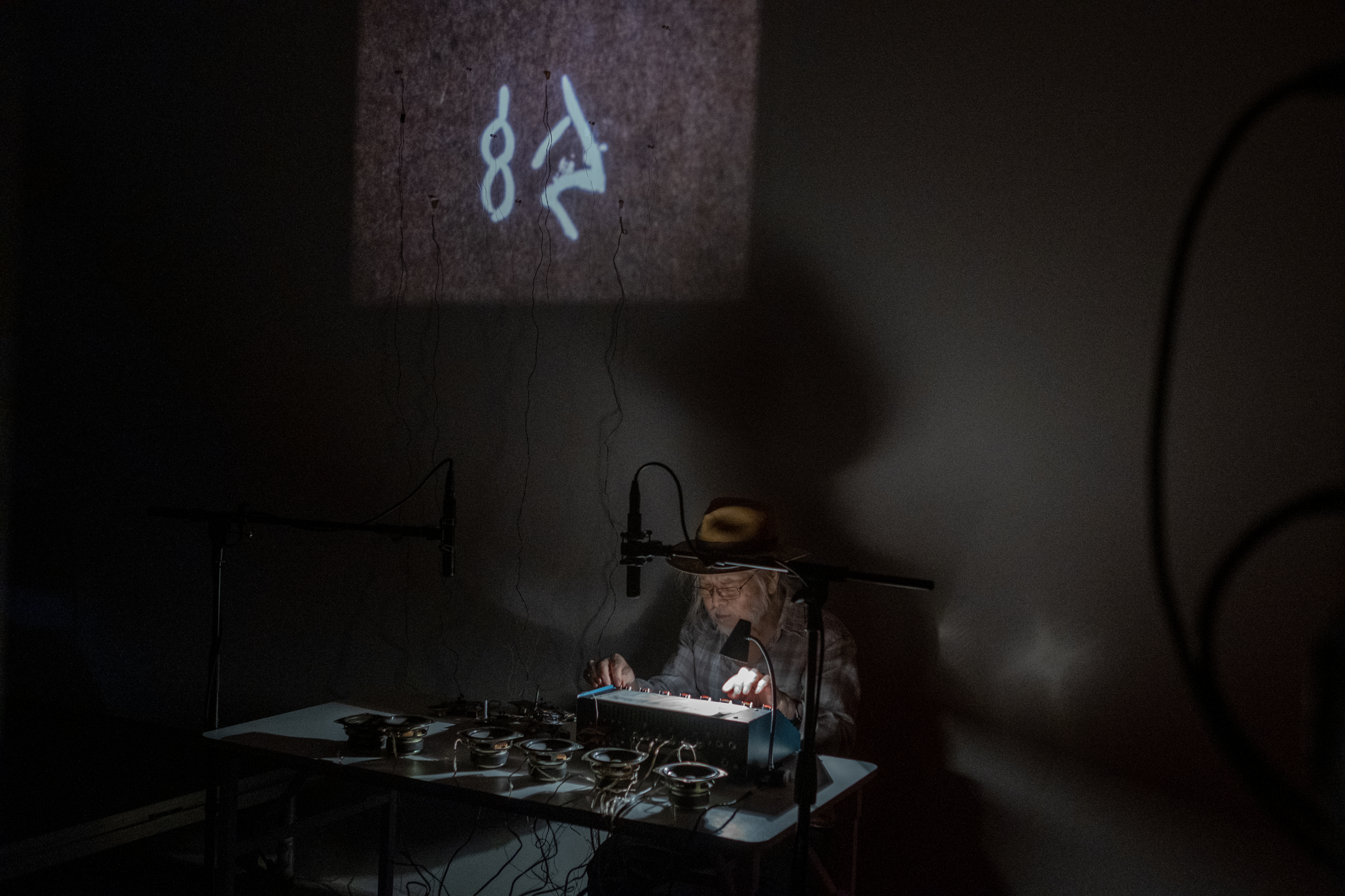 Color image of a darkly lit room. A figure sits behind a white table filled with audio equipment and two standing microphones are placed above the table. Light sensors are attached to the wall behind the figure where a projection of images plays.
