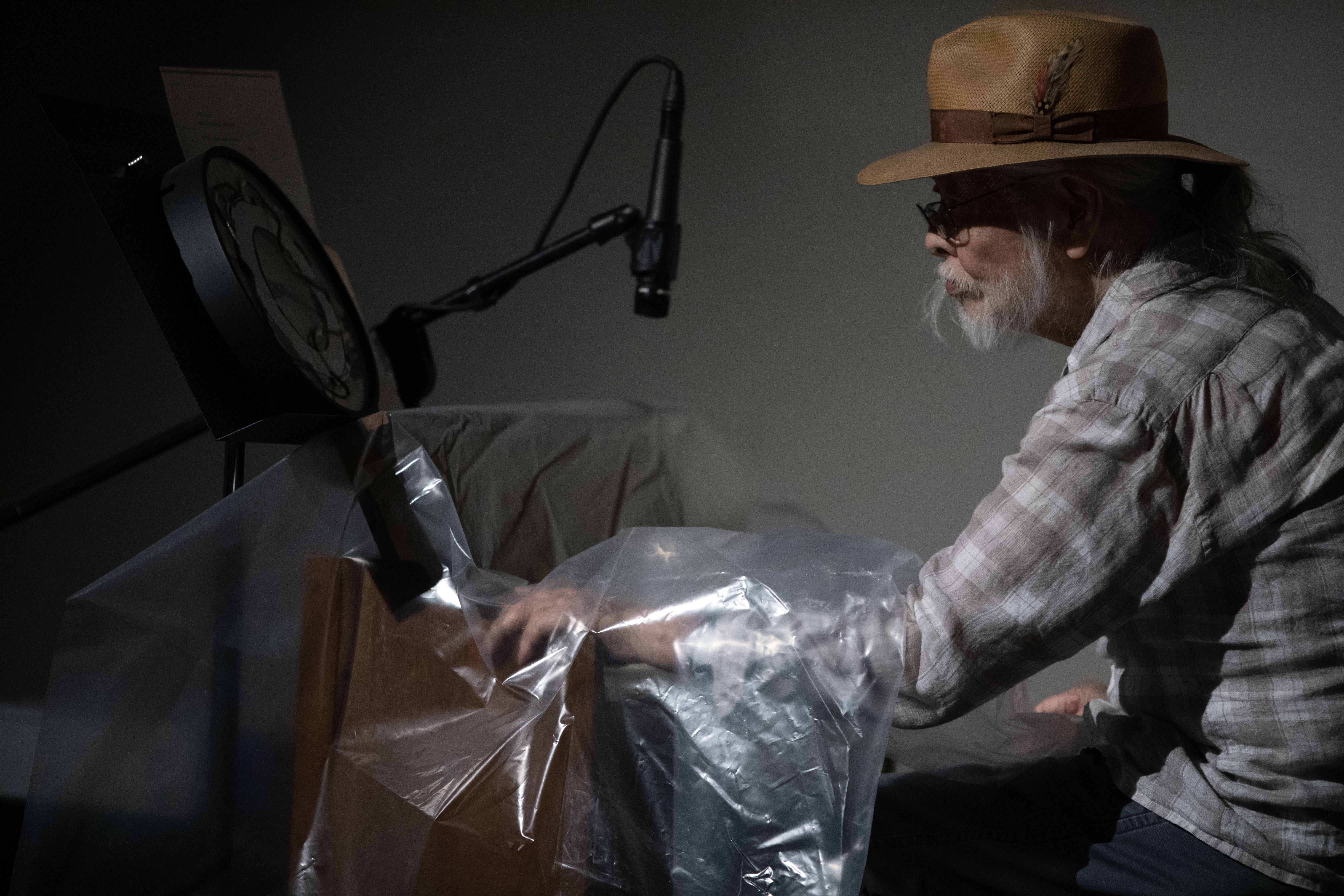 Color image of a figure in profile, seated behind a reed organ covered in a sheet of clear plastic. A clock sits on a music stand behind the organ. The figure reaches their arm under the plastic and holds the arm of the organ.