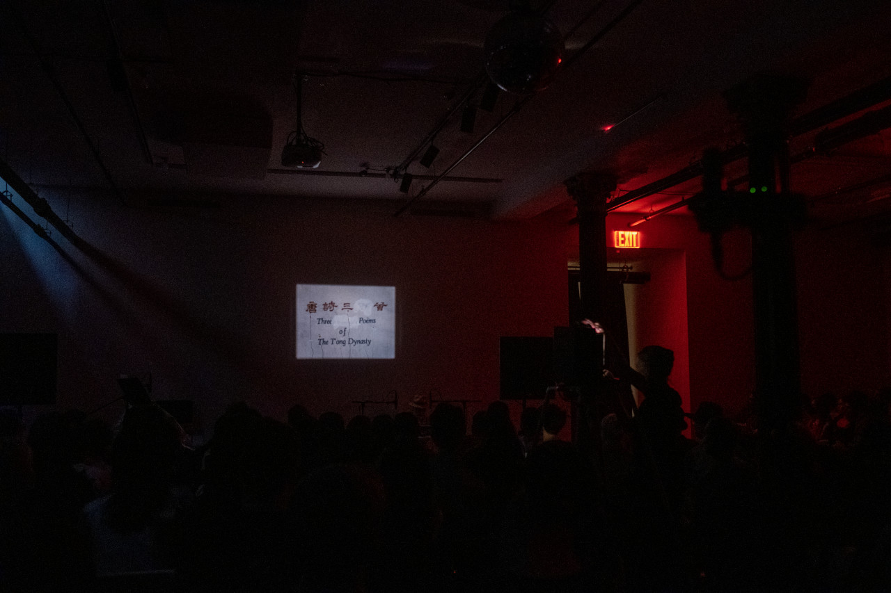 Color image of a dark basement room. On the back wall is projected an image with Chinese characters, and English characters that read "Three Poems of The Tong Dynasty."