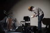 Color image of a figure standing on a black stage, with a spotlight placed on them. They are hunched over a black suitcase on the floor, and holding a small radio in their hands.