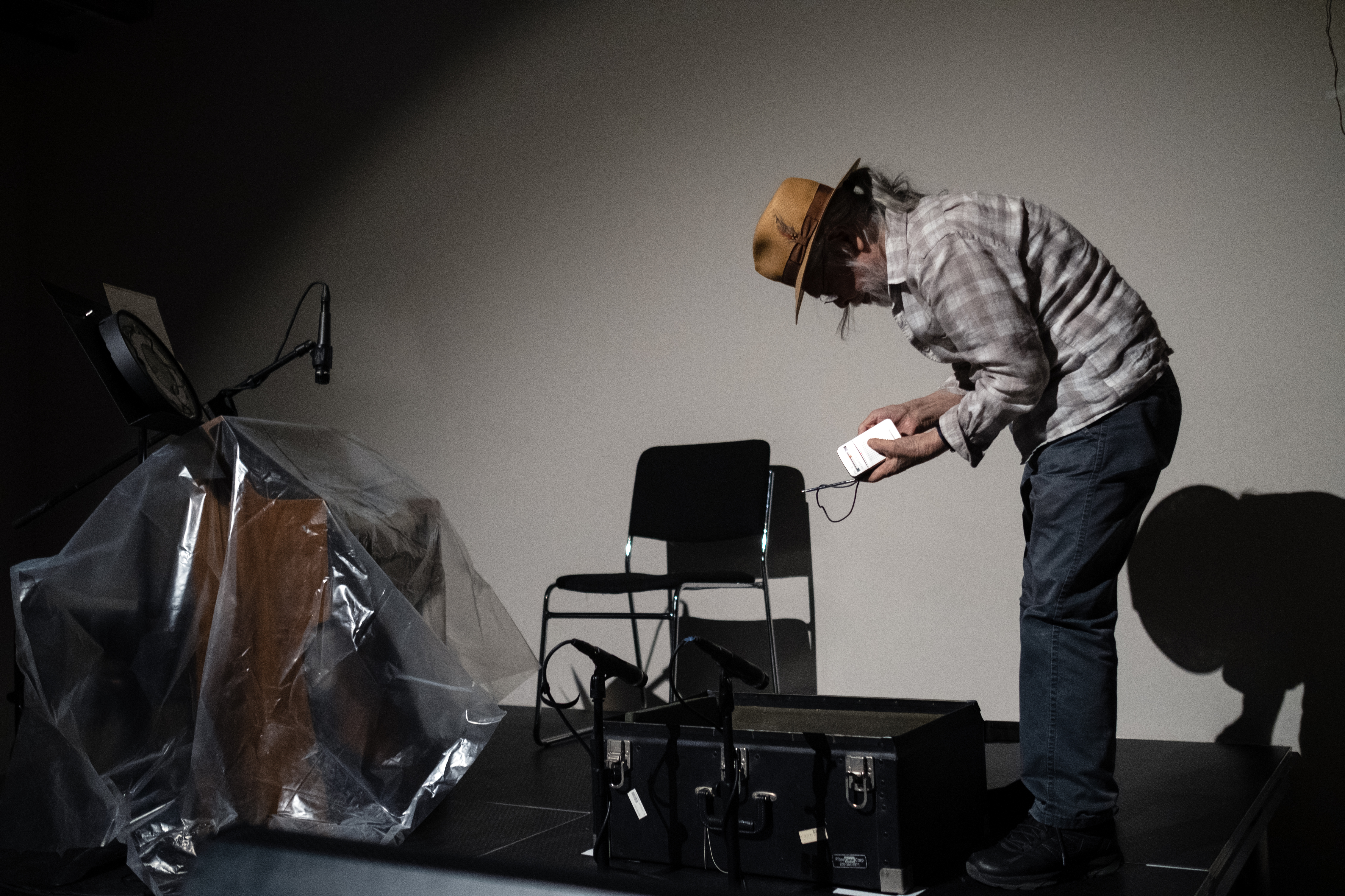 Color image of a figure standing on a black stage, with a spotlight placed on them. They are hunched over a black suitcase on the floor, and holding a small radio in their hands.