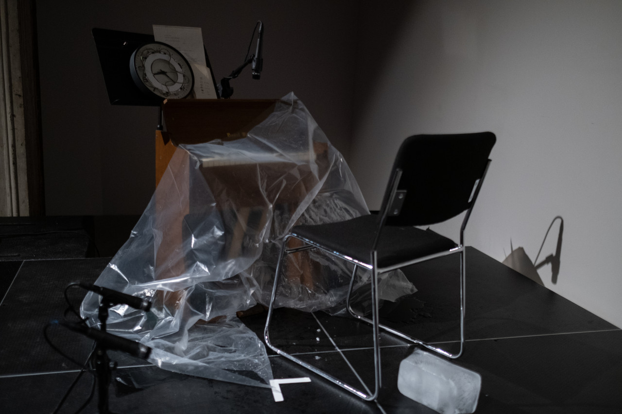 Color image of a reed organ with a sheet of clear plastic falling off the organ, and a block of ice fallen on its side on the floor of a black stage.