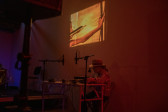 Color photograph of a figure seated behind a white desk filled with audio equipment. On the wall behind is a projected illustation of a figure holding a bow and arrow. The room is bathed in an orange/red light.