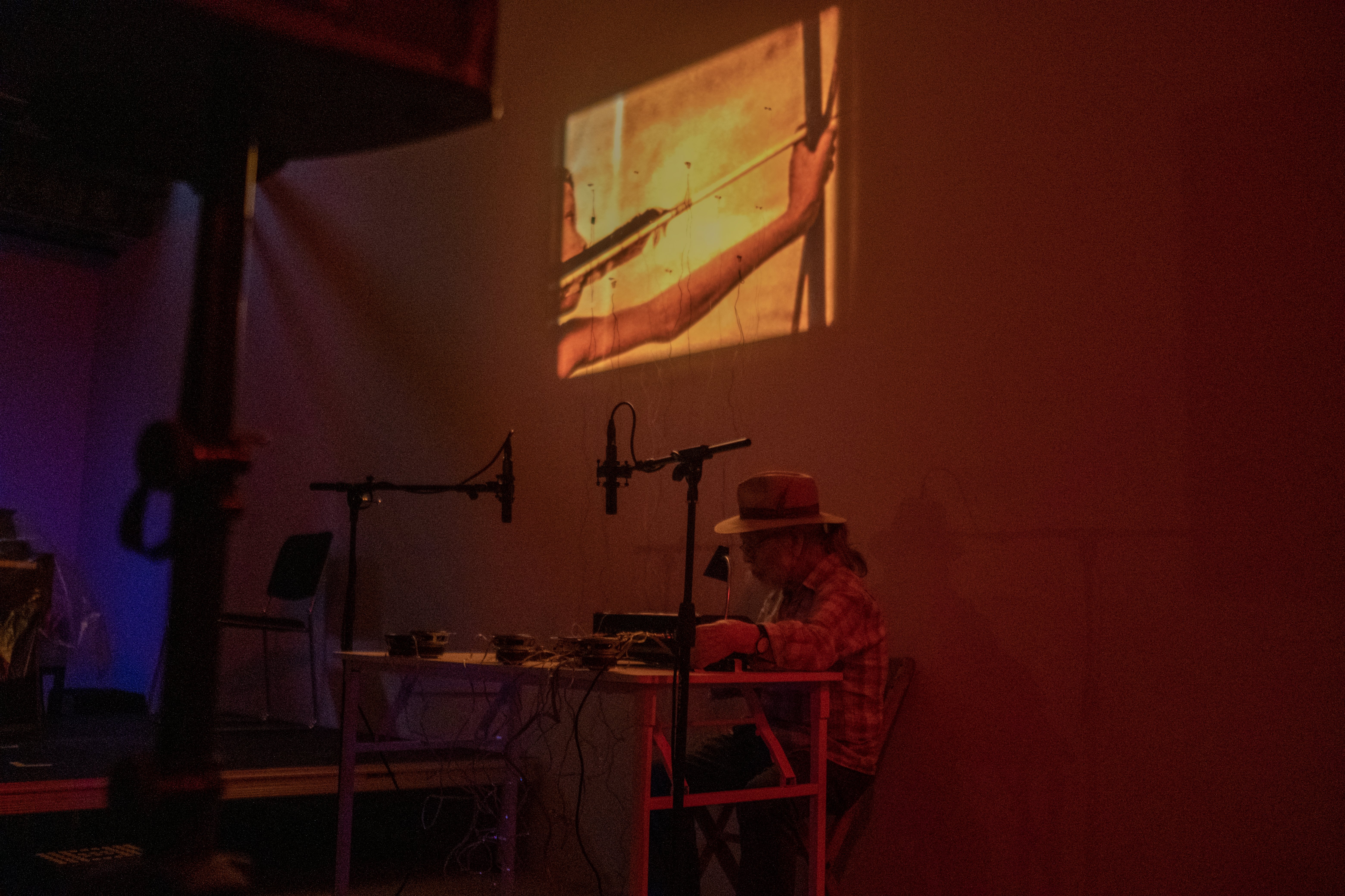 Color photograph of a figure seated behind a white desk filled with audio equipment. On the wall behind is a projected illustation of a figure holding a bow and arrow. The room is bathed in an orange/red light.