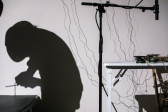 Color image of the silhouette of a figure on the right, they are bent over with a small radio in their hands. To the left is a standing microphone placed over speakers on a desk, with light sensors dangling on the wall behind.