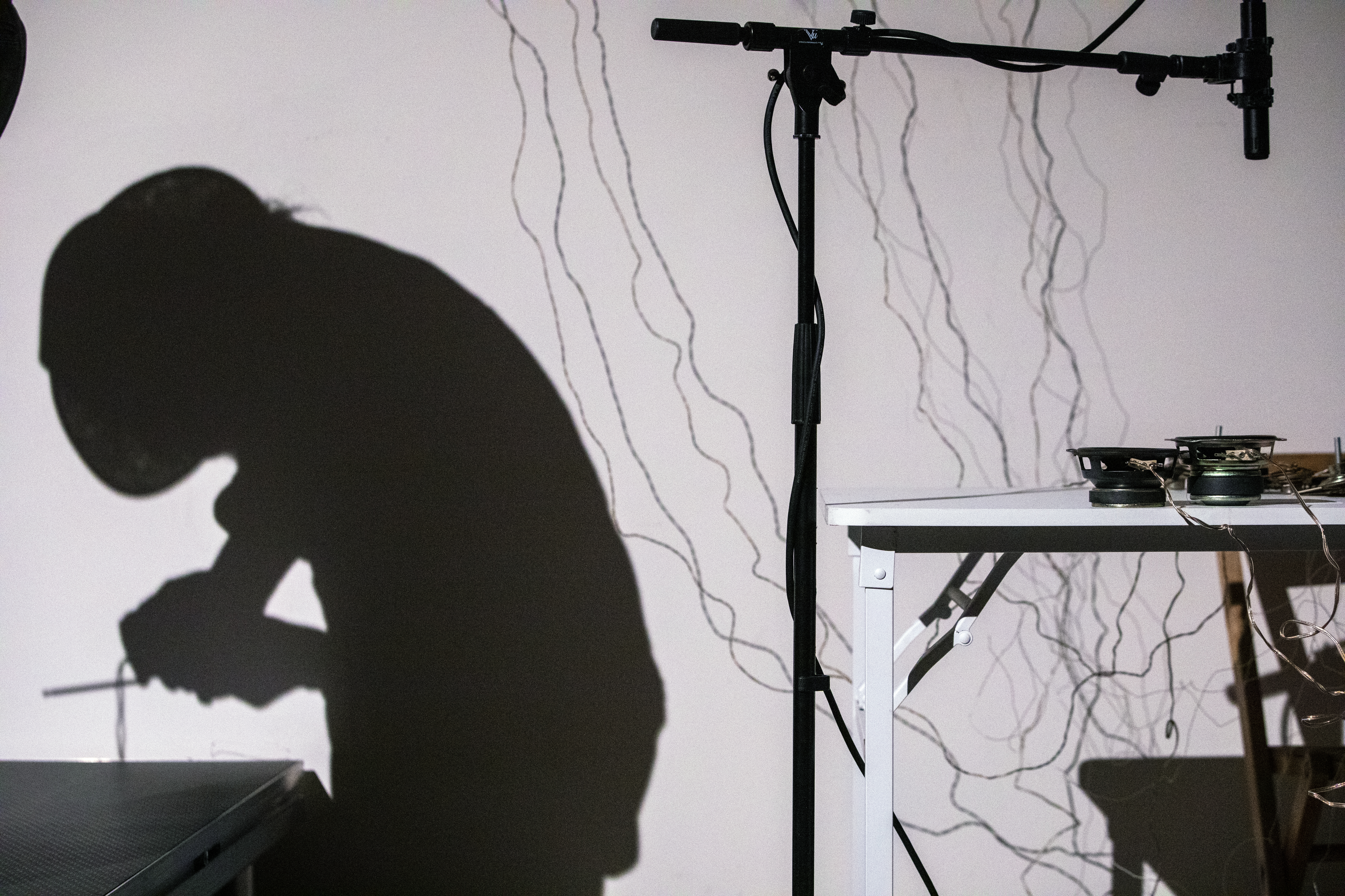 Color image of the silhouette of a figure on the right, they are bent over with a small radio in their hands. To the left is a standing microphone placed over speakers on a desk, with light sensors dangling on the wall behind.