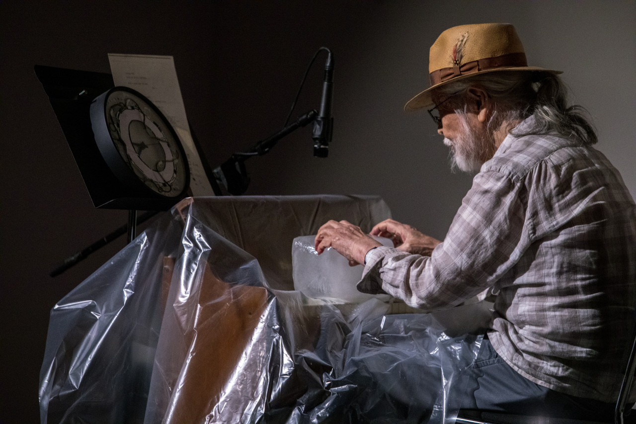 Color image of a figure sitting behind a reed organ covered in a sheet of clear plastic. A clock sits on a music stannd with instructions behind the organ. The figure positions their hands on a large block of ice that sits on top of the keys of the organ.