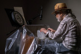 Color image of a figure sitting behind a reed organ covered in a sheet of clear plastic. A clock sits on a music stannd with instructions behind the organ. The figure positions their hands on a large block of ice that sits on top of the keys of the organ.