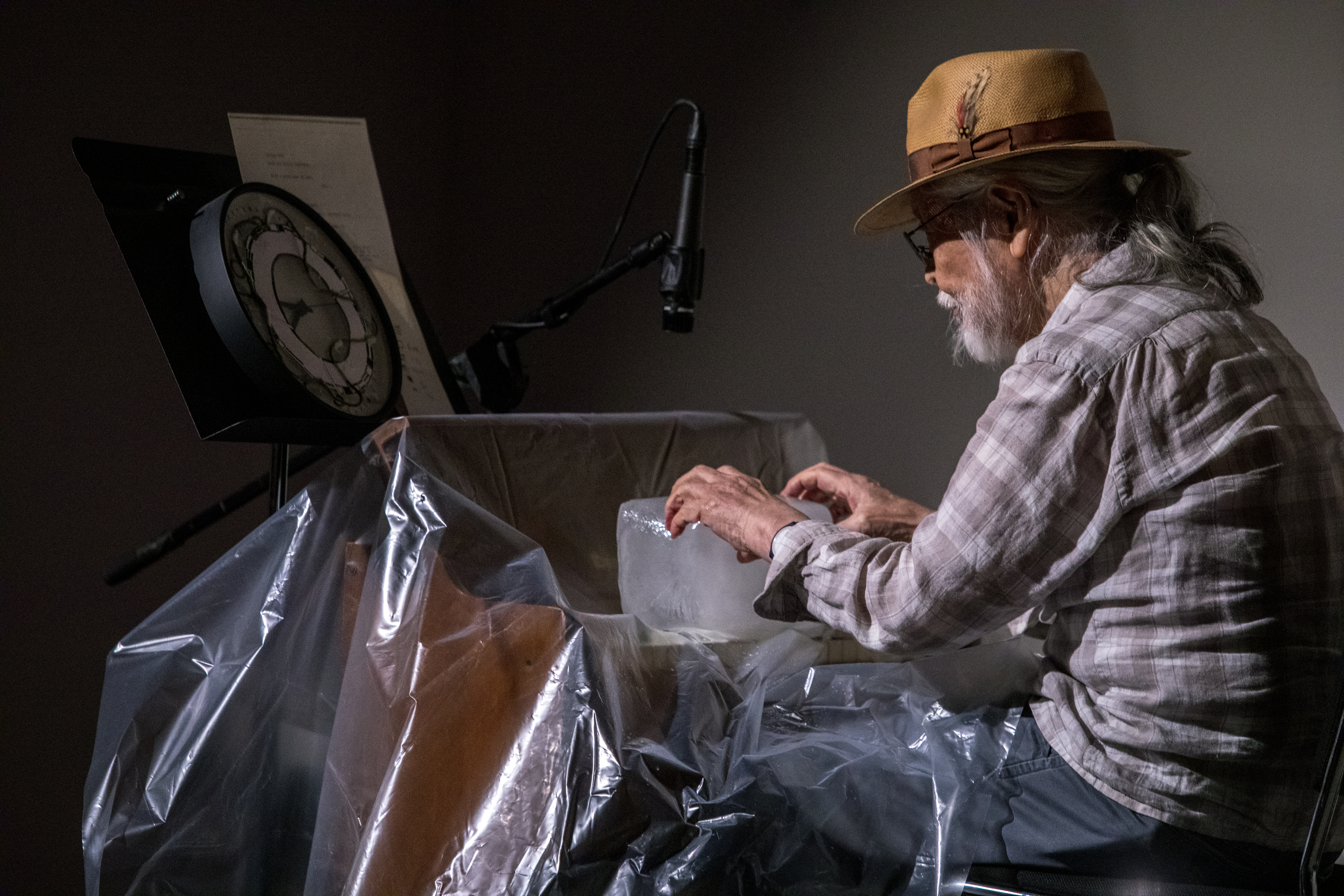 Color image of a figure sitting behind a reed organ covered in a sheet of clear plastic. A clock sits on a music stannd with instructions behind the organ. The figure positions their hands on a large block of ice that sits on top of the keys of the organ.