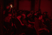Color image of a seated audience all facing the right of the room. A figure in the back middle is standing and operating a film projector. The room is bathed in red light.
