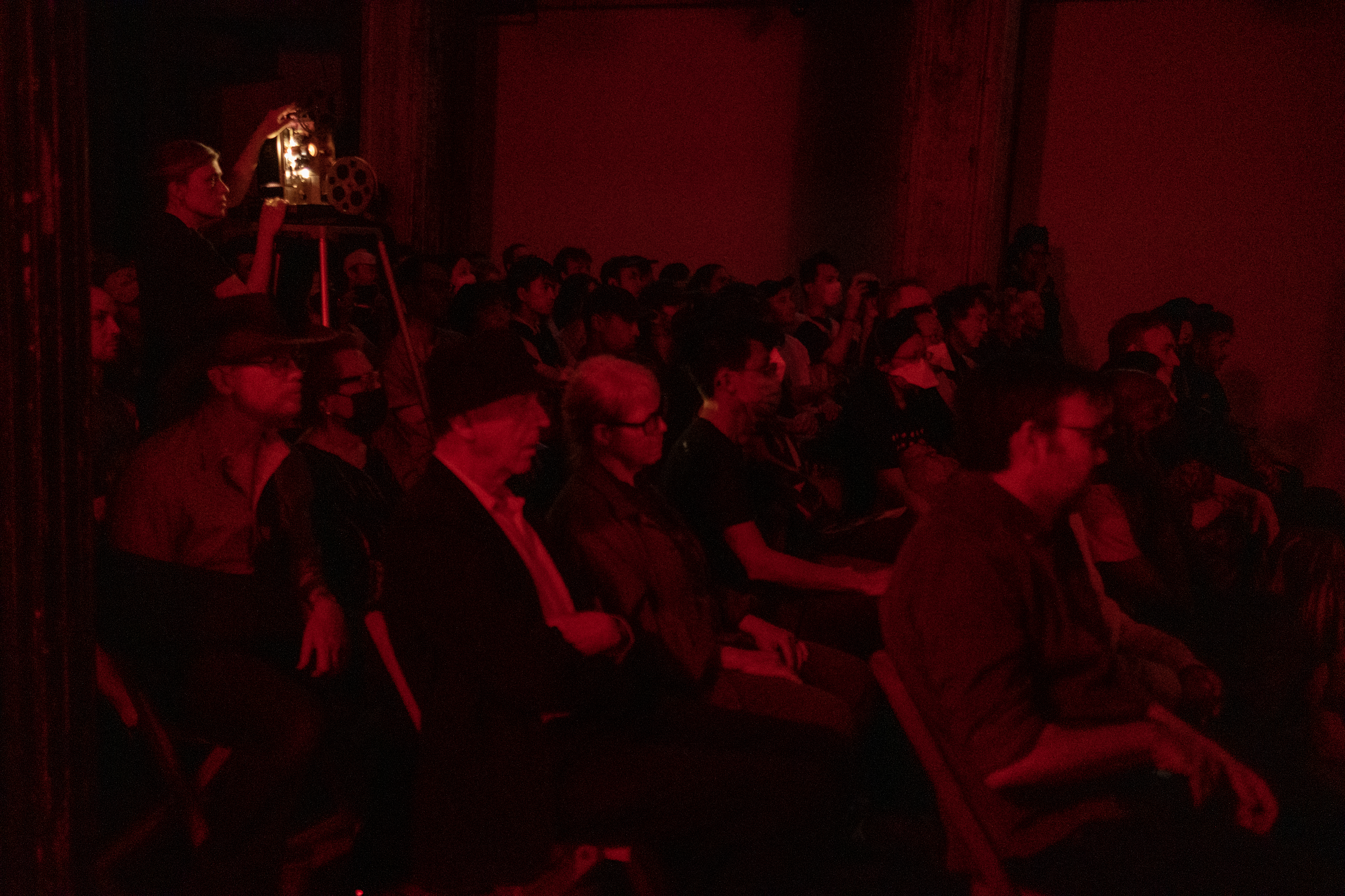 Color image of a seated audience all facing the right of the room. A figure in the back middle is standing and operating a film projector. The room is bathed in red light.