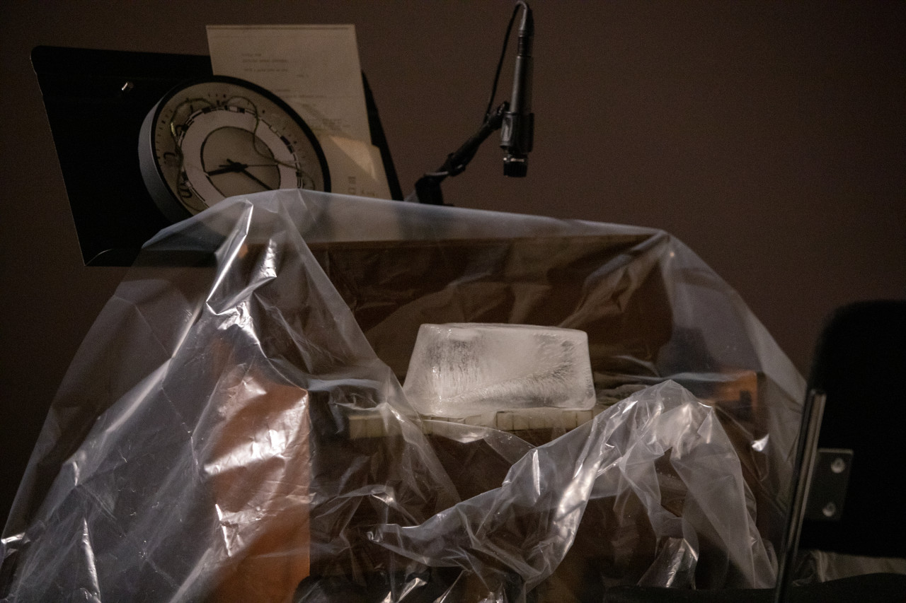 Color image of an ice block sitting on top of the keys of a reed organ. The reedorgan is covered in a sheet of clear plastic. A clock and instructions sit on a music stand behind the organ.