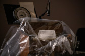 Color image of an ice block sitting on top of the keys of a reed organ. The reedorgan is covered in a sheet of clear plastic. A clock and instructions sit on a music stand behind the organ.
