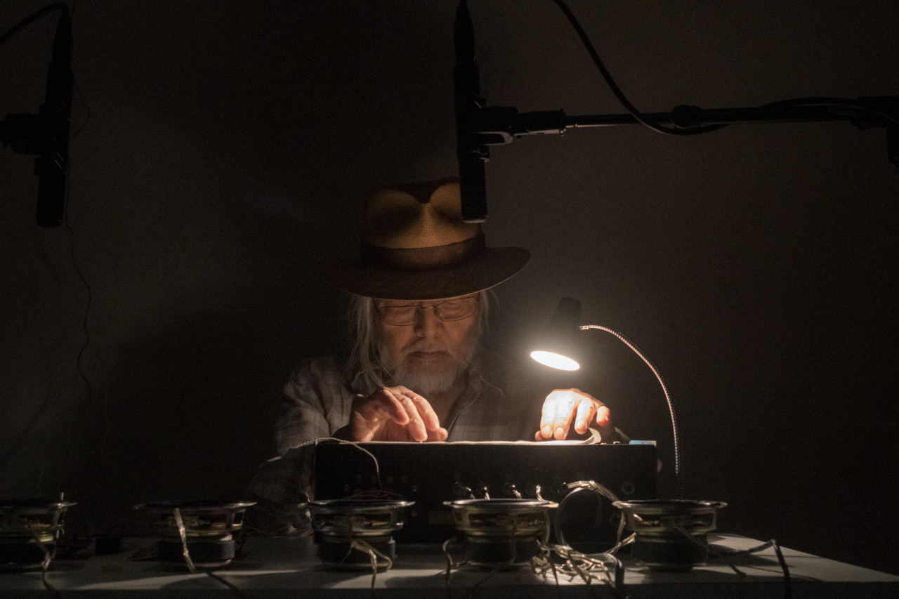 Color image of a figure sitting behind a white desk, manipulating an audio machine with their hands. A lamp illuminates the machines controls and speakers are placed in front of the machine.