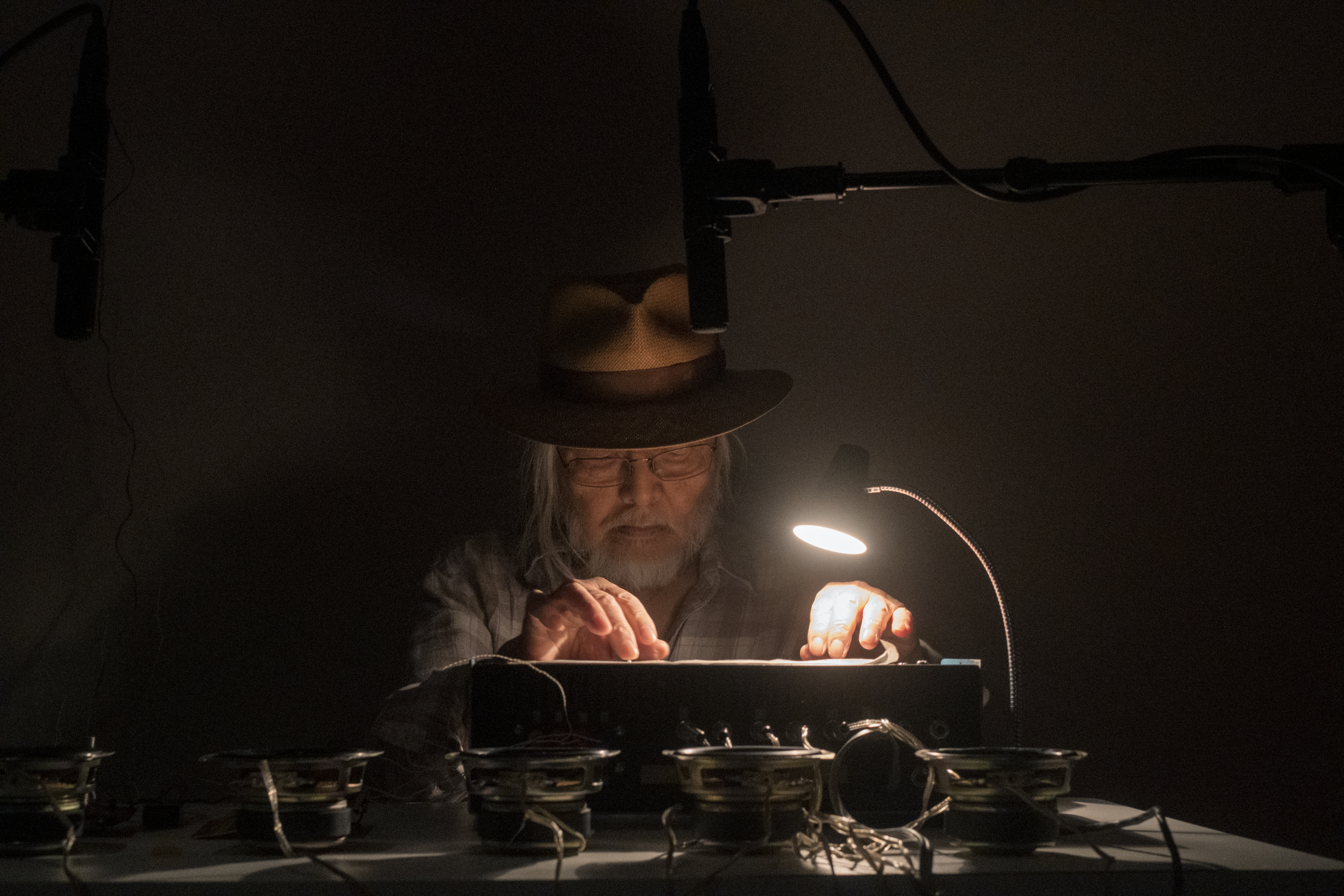 Color image of a figure sitting behind a white desk, manipulating an audio machine with their hands. A lamp illuminates the machines controls and speakers are placed in front of the machine.