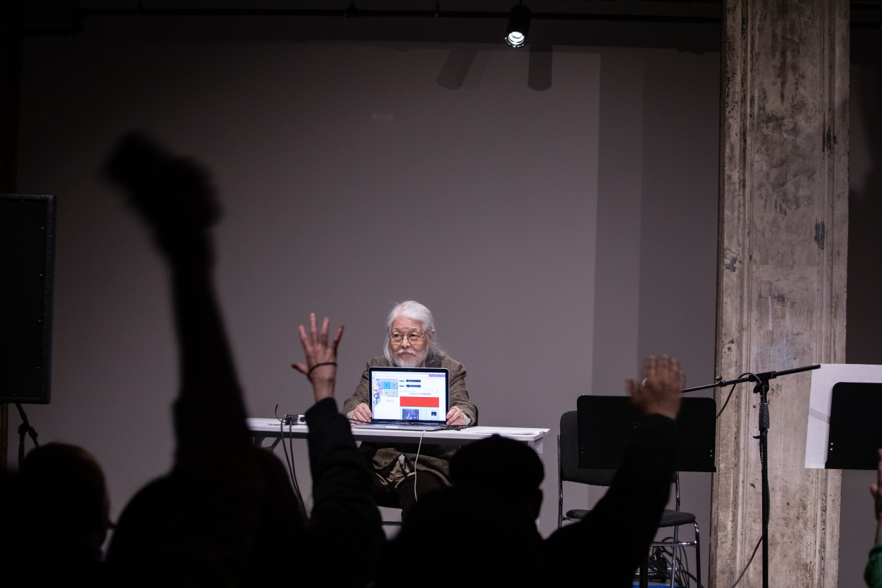 Color image of a figure seated behind a white desk on a stage. They face out into the audience, and controls a laptop that also faces out into the audience. Members of the audience wave their arms in the air so the laptop will capture their movement.
