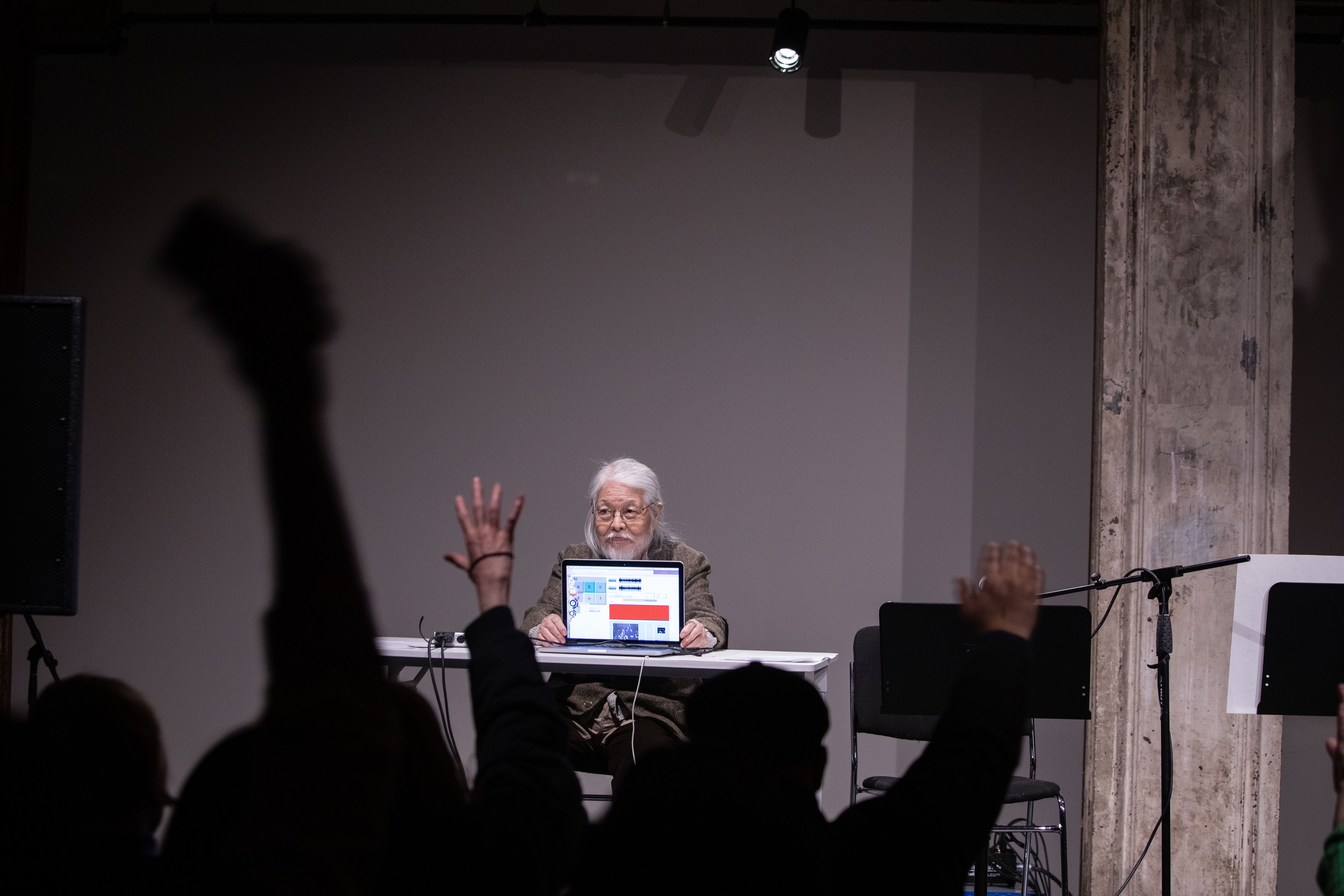 Color image of a figure seated behind a white desk on a stage. They face out into the audience, and controls a laptop that also faces out into the audience. Members of the audience wave their arms in the air so the laptop will capture their movement.