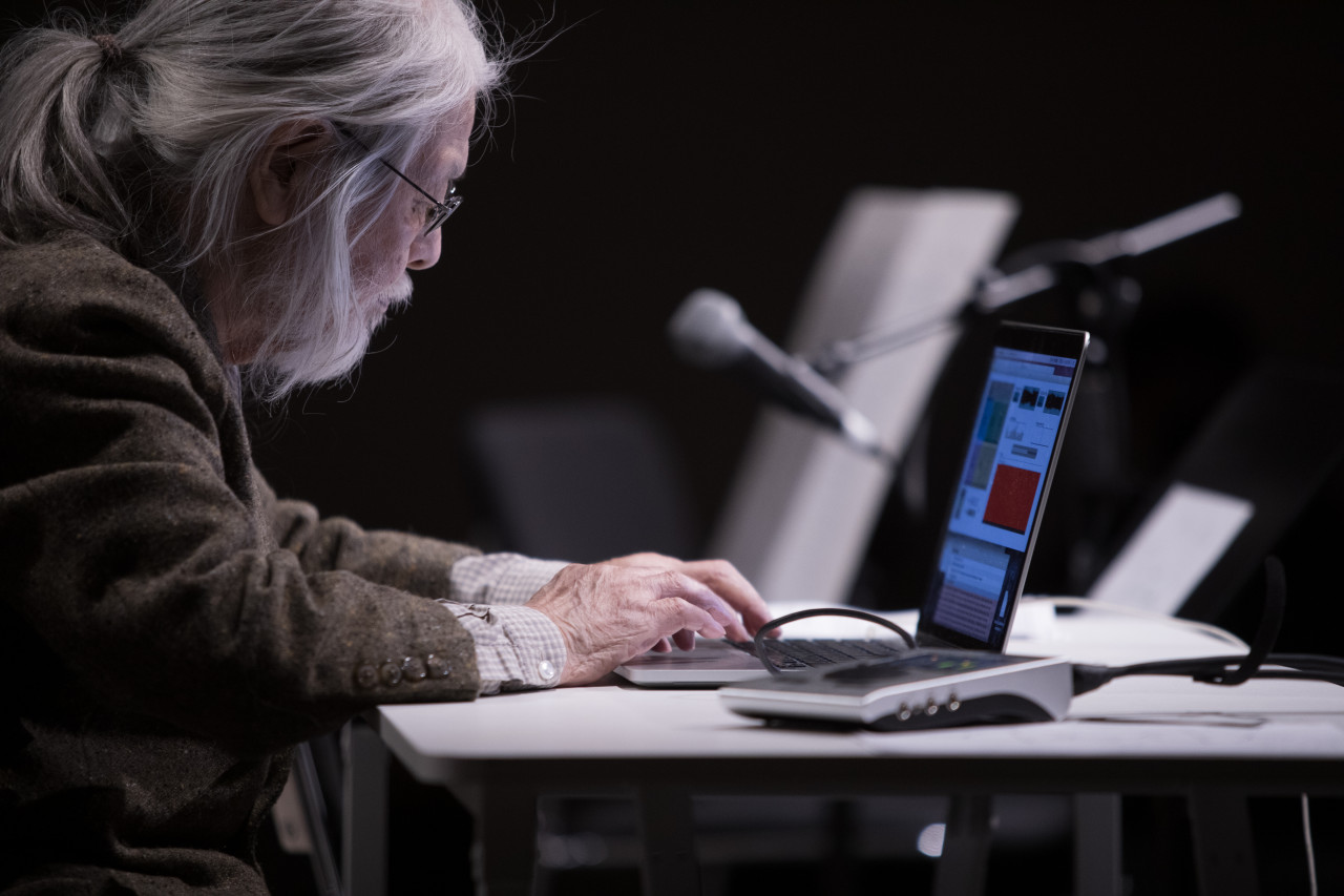 Color image of a figure seated behind a white desk. They are using a laptop, open to a computer program page.