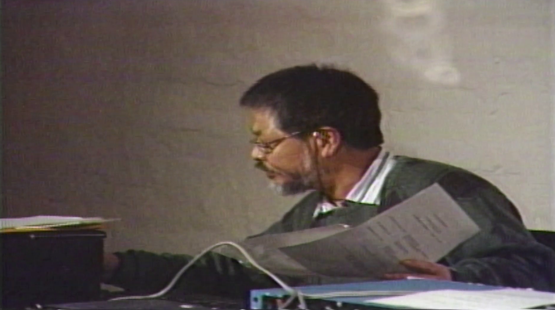 Color image of a figure looking to the left and leaning over a table filled with audio equipment. The figure holds sheets of paper with instructions, and manipulates an audio machine.