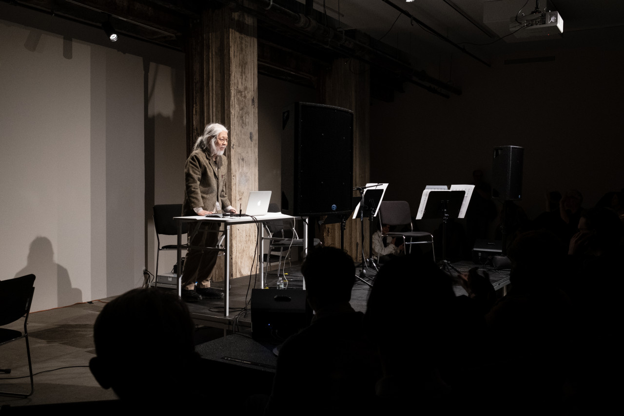 Color image of a figure performing on a black stage in front of an audience. The figure on stage stands behind a white desk with a laptop and other audio equipment on it.