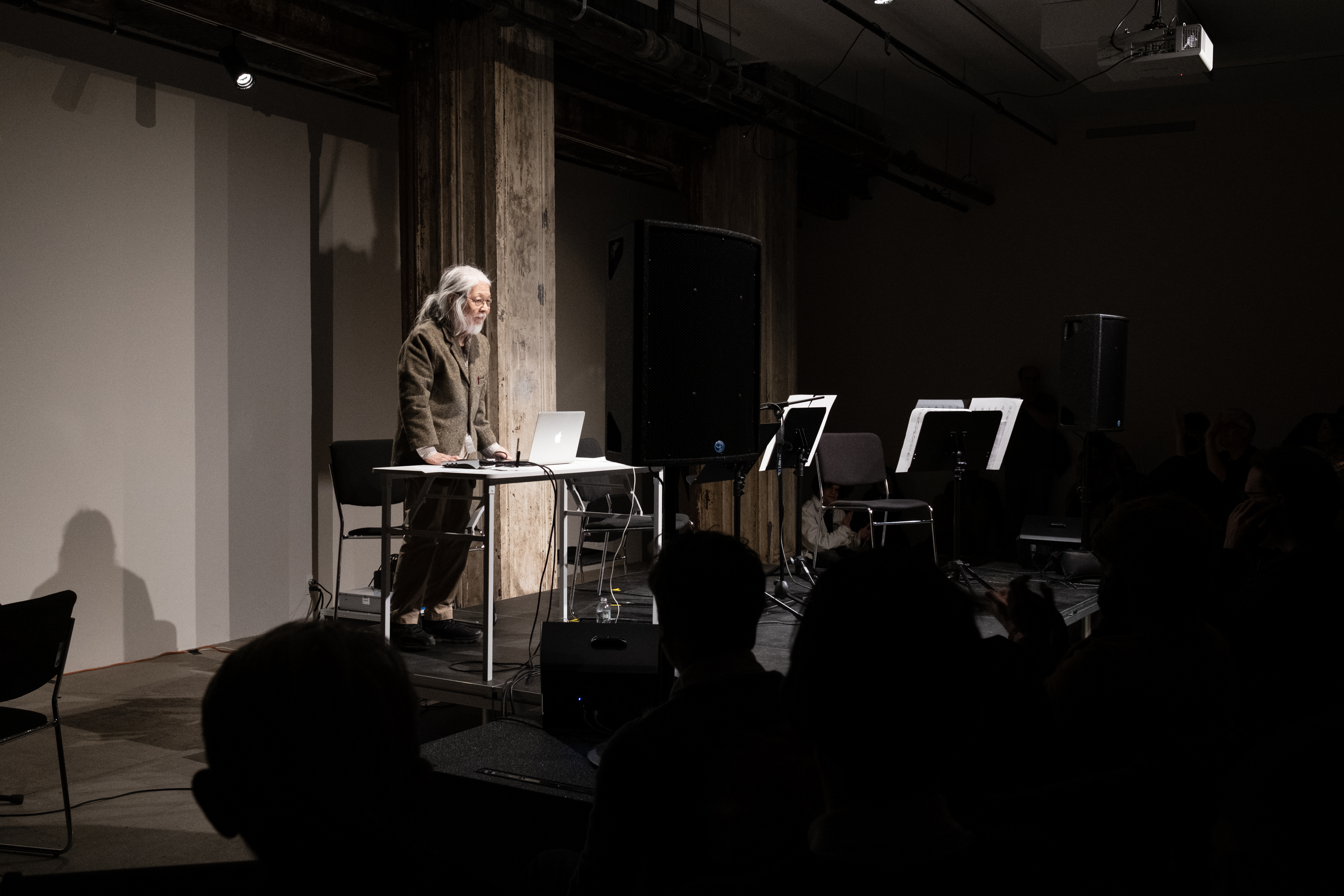 Color image of a figure performing on a black stage in front of an audience. The figure on stage stands behind a white desk with a laptop and other audio equipment on it.