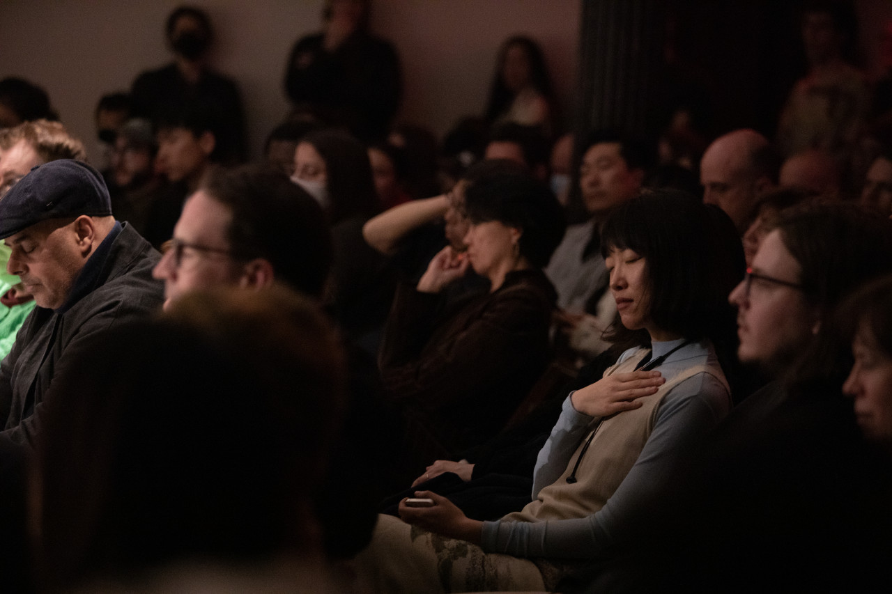 Color image of an audience watching a performance. In the middle of the crowd, a figure in a blue and white dress holds their hand to their chest with their eyes closed, listening to the performance.