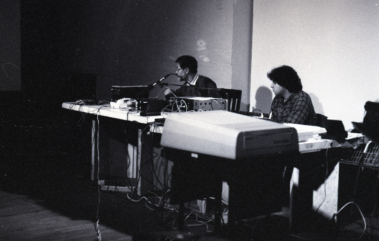 Black-and-white photograph of two figures sitting behind tables filled with audio equipment. They are focused on manipulating the audio machines in front of them.