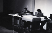 Black-and-white photograph of two figures sitting behind tables filled with audio equipment. They are focused on manipulating the audio machines in front of them.