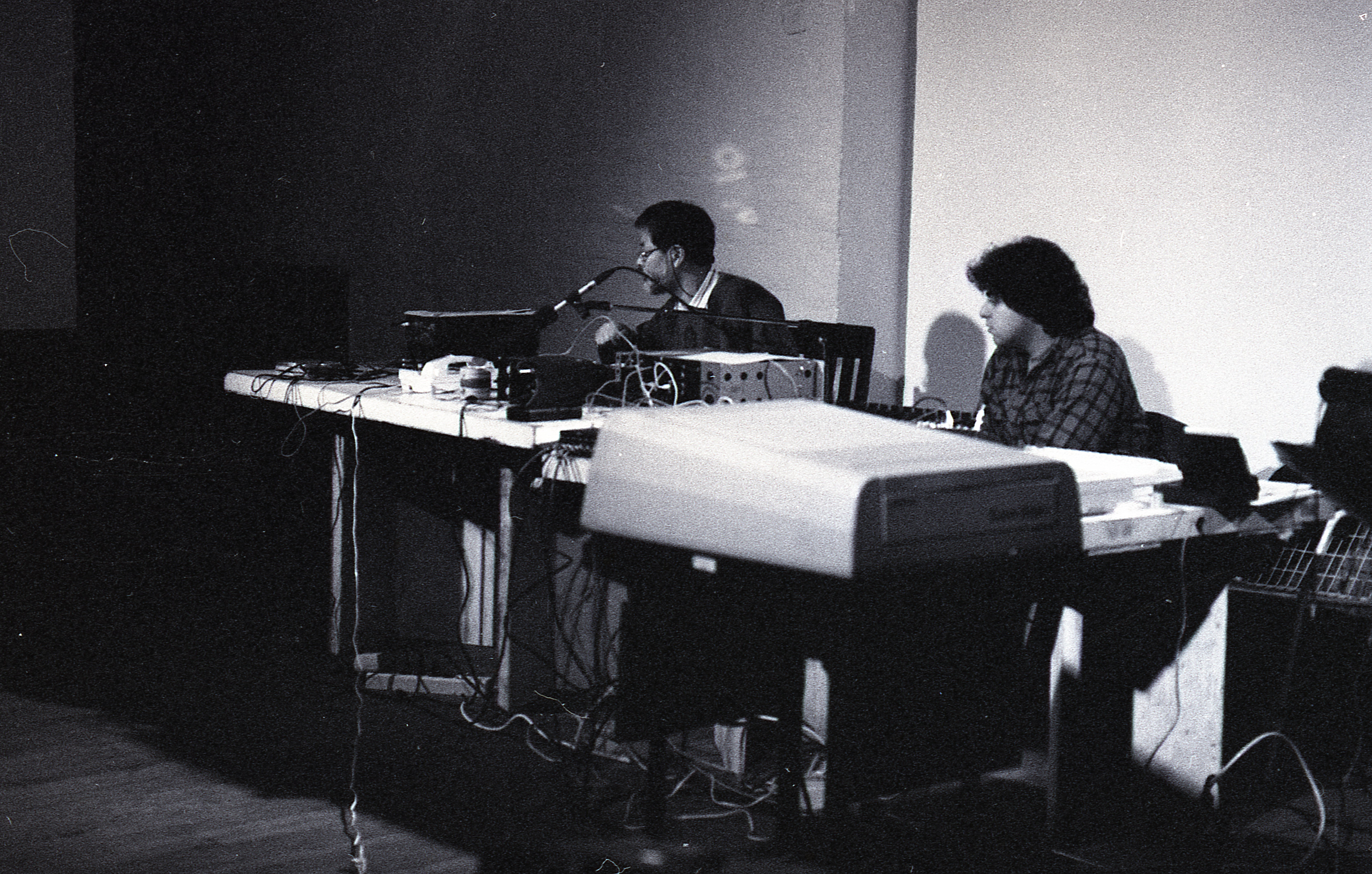 Black-and-white photograph of two figures sitting behind tables filled with audio equipment. They are focused on manipulating the audio machines in front of them.