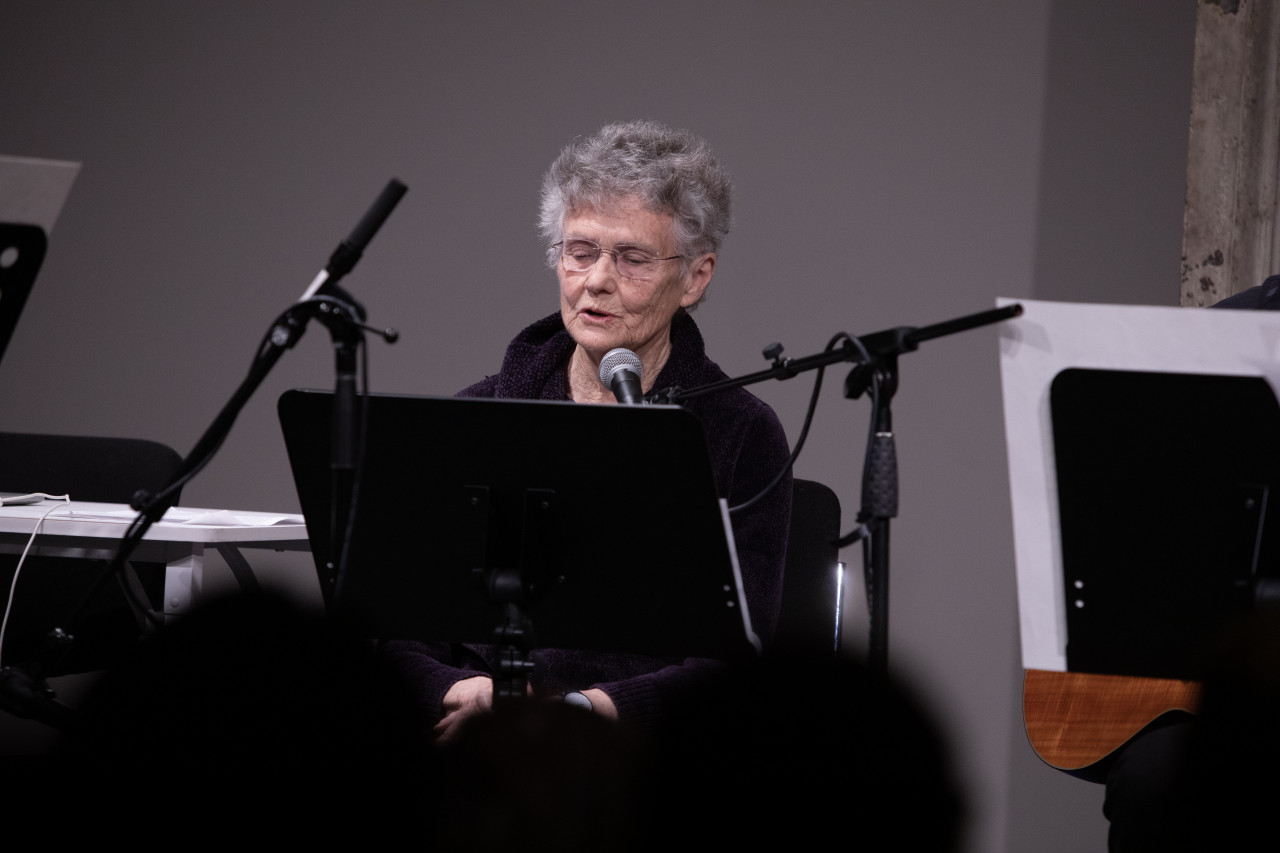 Color image of a figure reading into a microphone, sitting behind a music stand.