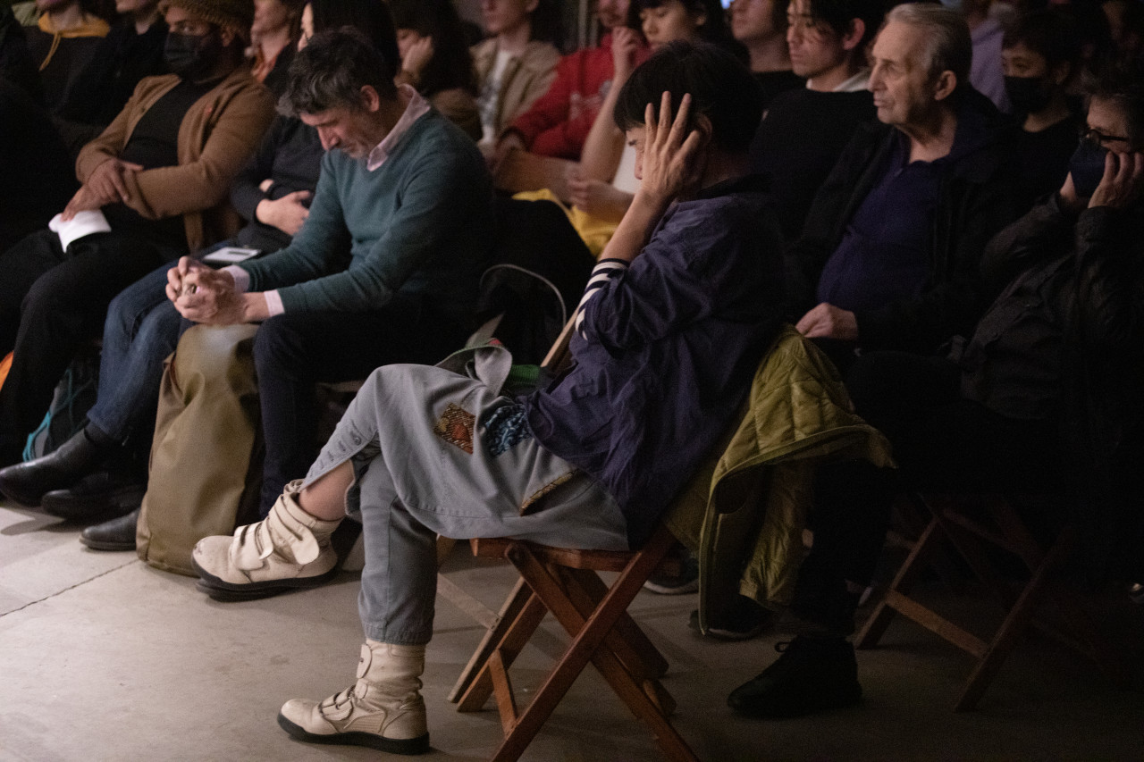 Color image of an audience listening to a performance. In the middle of the image, a figure sitting on a wooden chair holds their hands over their ears and looks downward.
