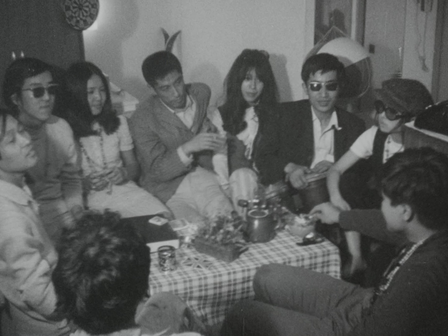Black-and-white image of nine figures sitting around a small table. One figure is playing a hand drum while the others chat and smoke.