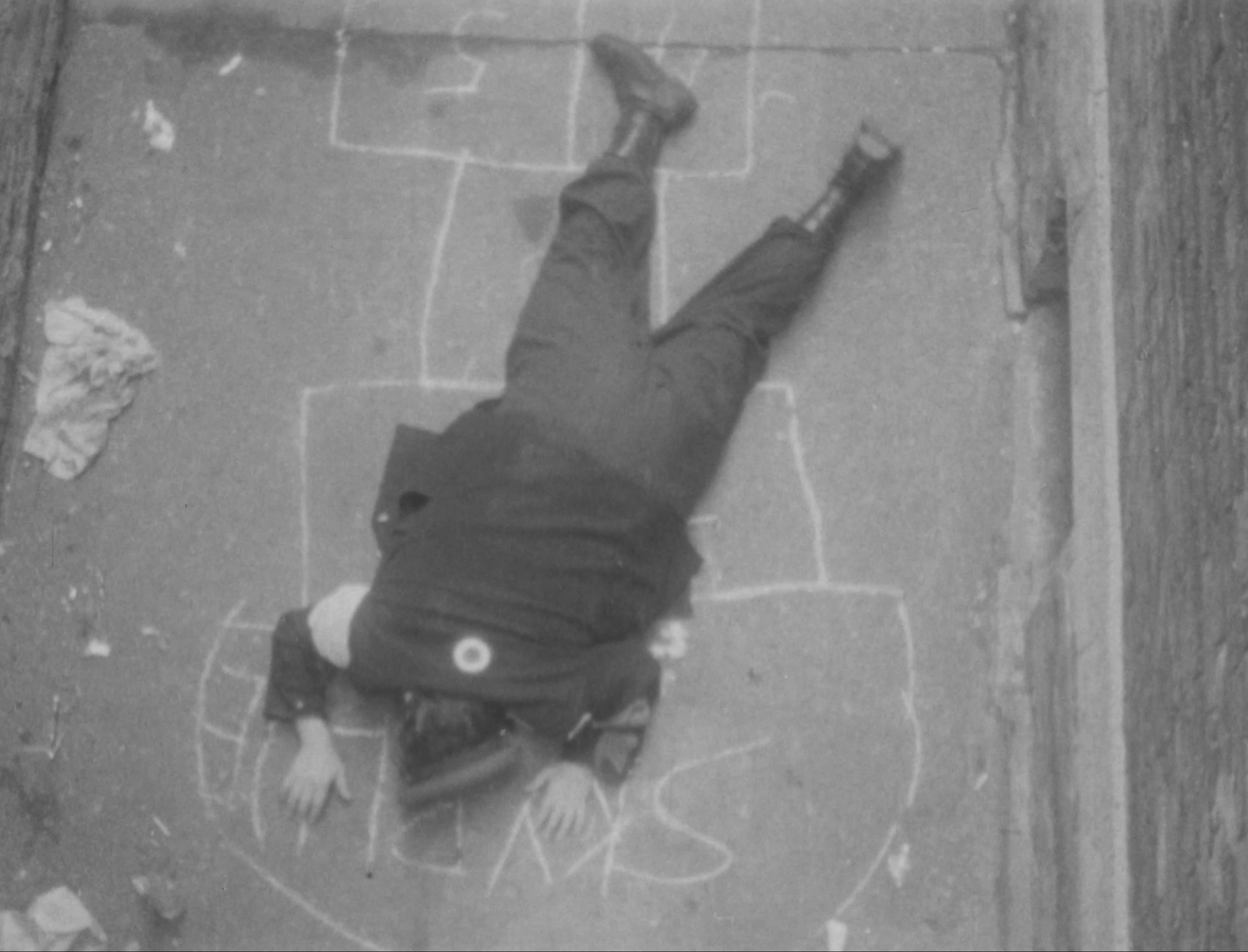 Black-and-white image of a figure in uniform laying face down on the street. A hopscotch pattern is drawn in white chalk on the ground beneath the figure.