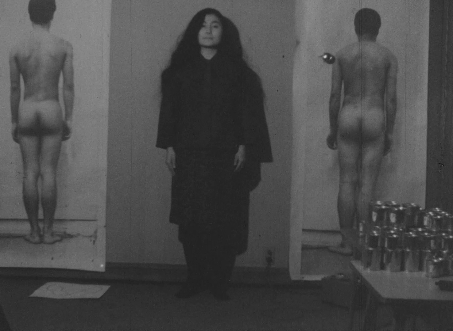 Black-and-white image of a figure in black standing straight against a wall. On either side of them are life-size printed images of a naked figure from behind.