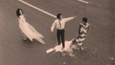 Sepia toned image of three figures standing in the middle of the street. The figure on the left wears a white dress that flows in the wind, while the middle figure wears a button down shirt and stands in a pool of white liquid spilled on the ground. The third figure on the right wears a striped cloth over their body.