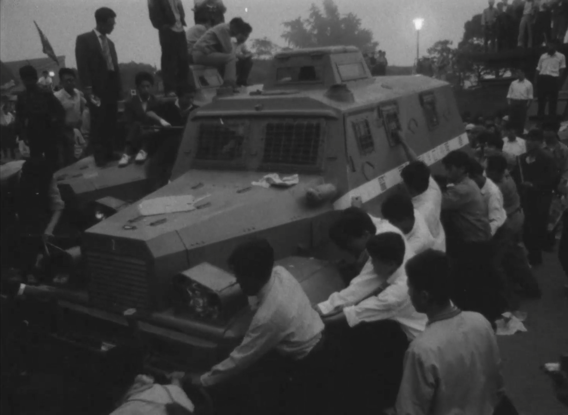 Black-and-white image of a group of figures in suits and white shirts surrounding a large military tank, they are grabbing the sides and trying to control the vehicle.