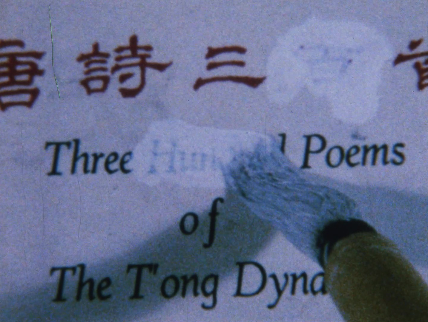 Color image of a white page with Chinese and English printed text on it. A brush with white paint is painting over the word 