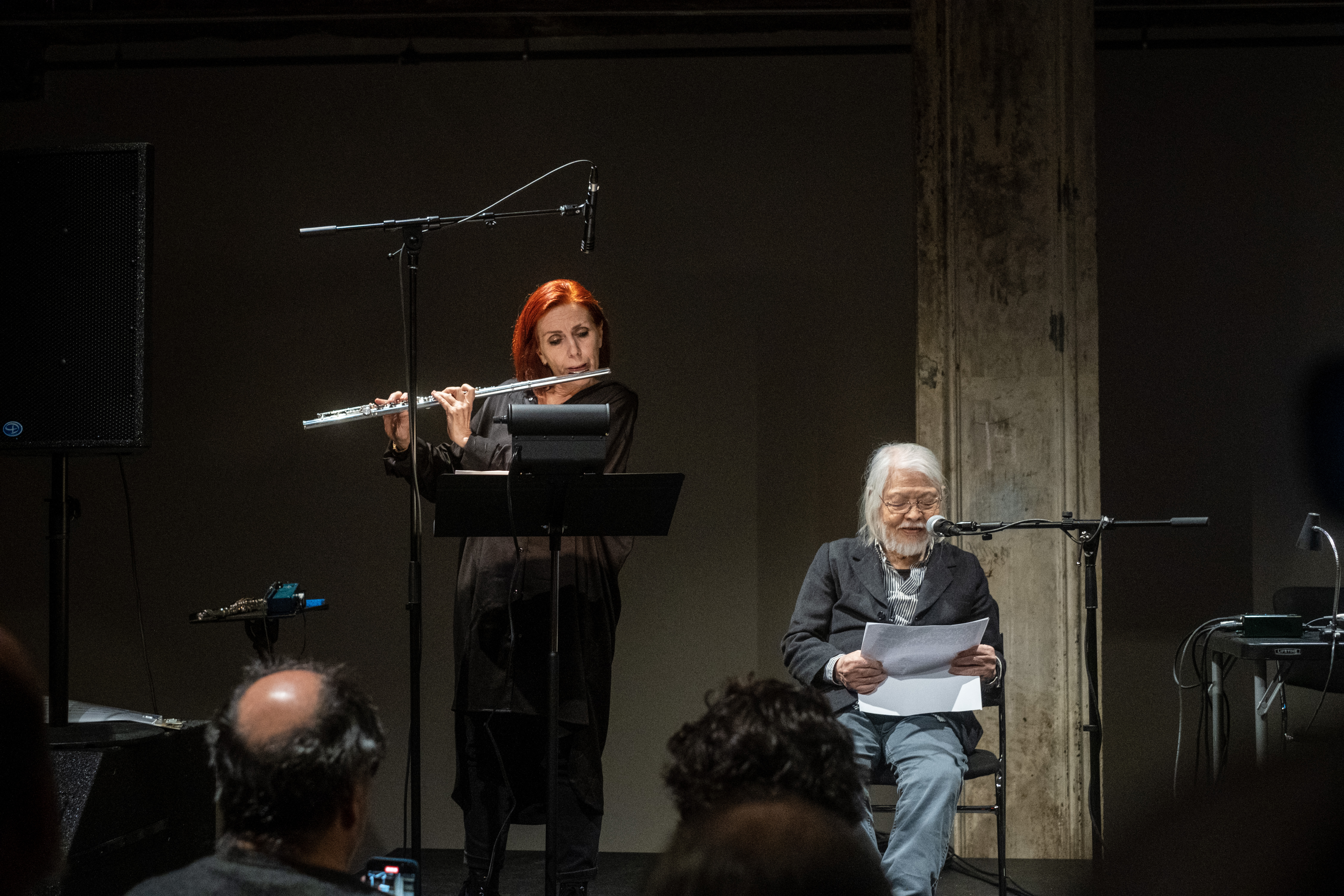 Two figures perform on stage. The figure on the right stands behind a music stand and plays the flute while the figure on the left sits behind a microphone and reads off a piece of paper.