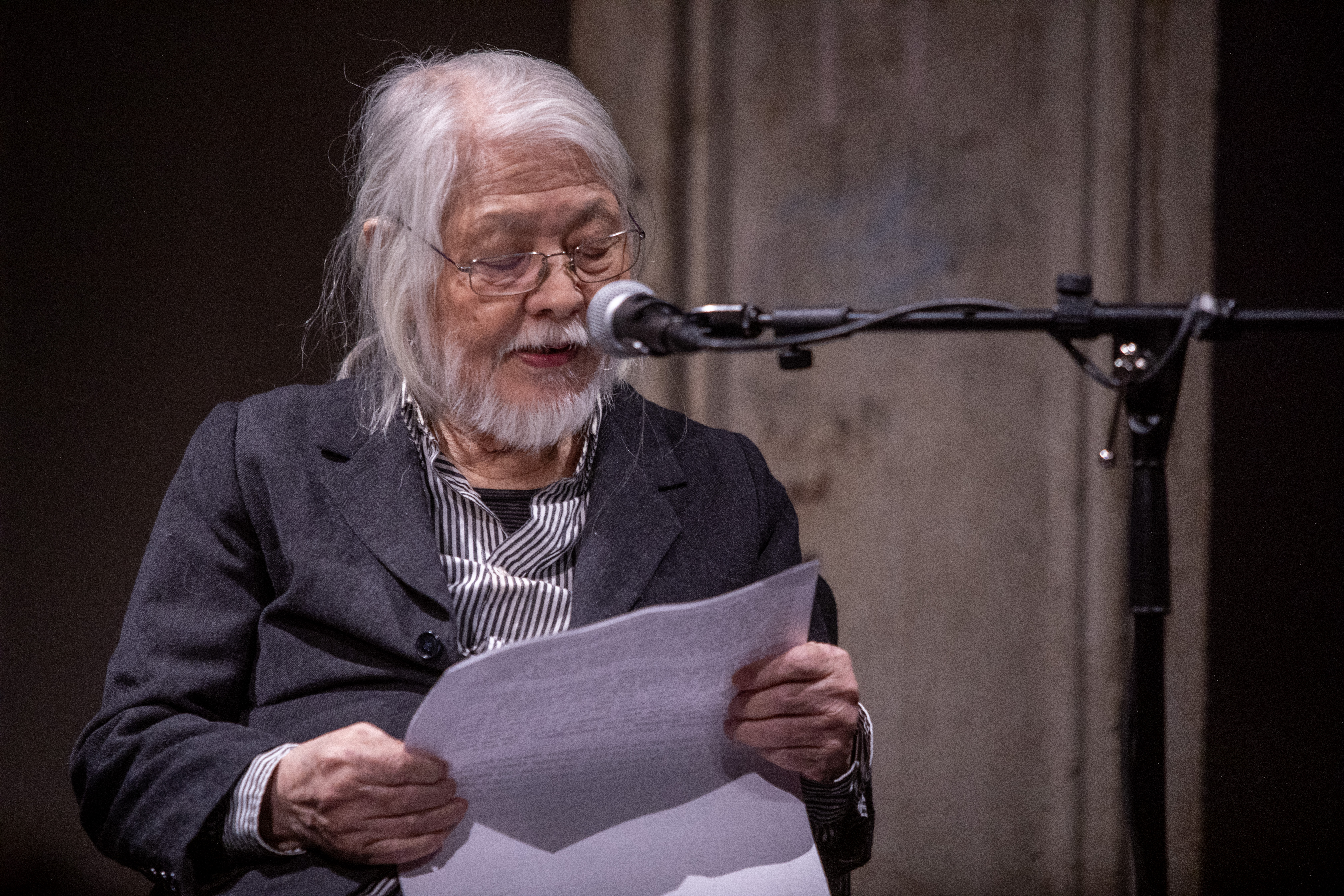 A figure sits in front of a microphone and reads a story off of a piece of paper.