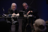 Two figures sit in front of a table filled with audio equipment including a commodore computer and flute-to-haiku converter.