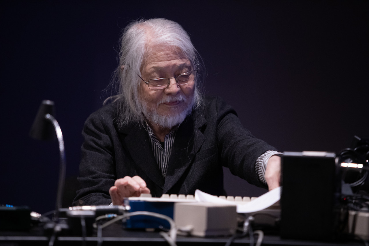 A figure sits in front of a table with audio equipment, including a commodore computer and flute-to-haiku converter.