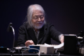 A figure sits in front of a table with audio equipment, including a commodore computer and flute-to-haiku converter.