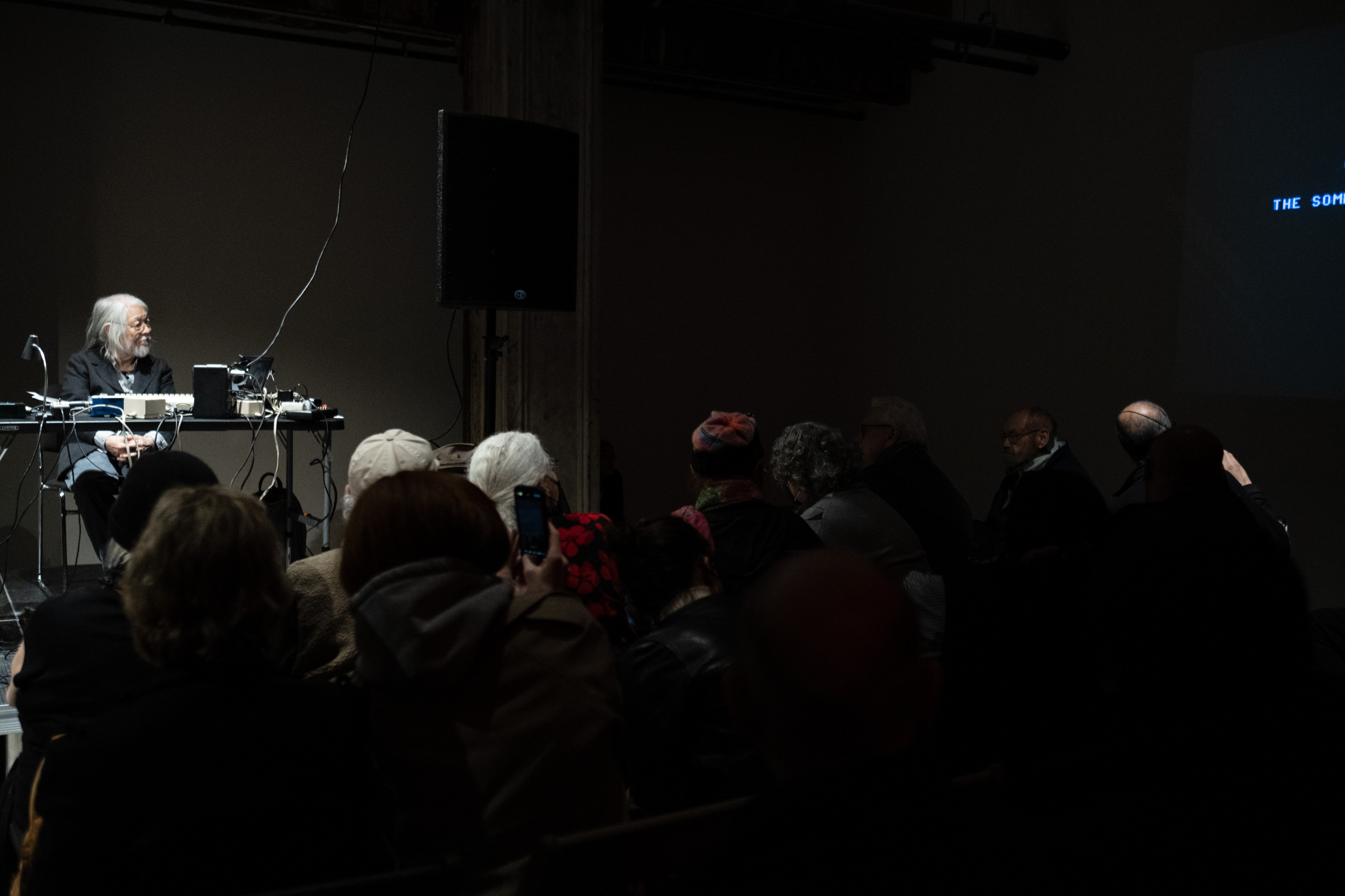 A figure sits on stage in front of a crowd and looks to the right as text is projected onto the right wall.