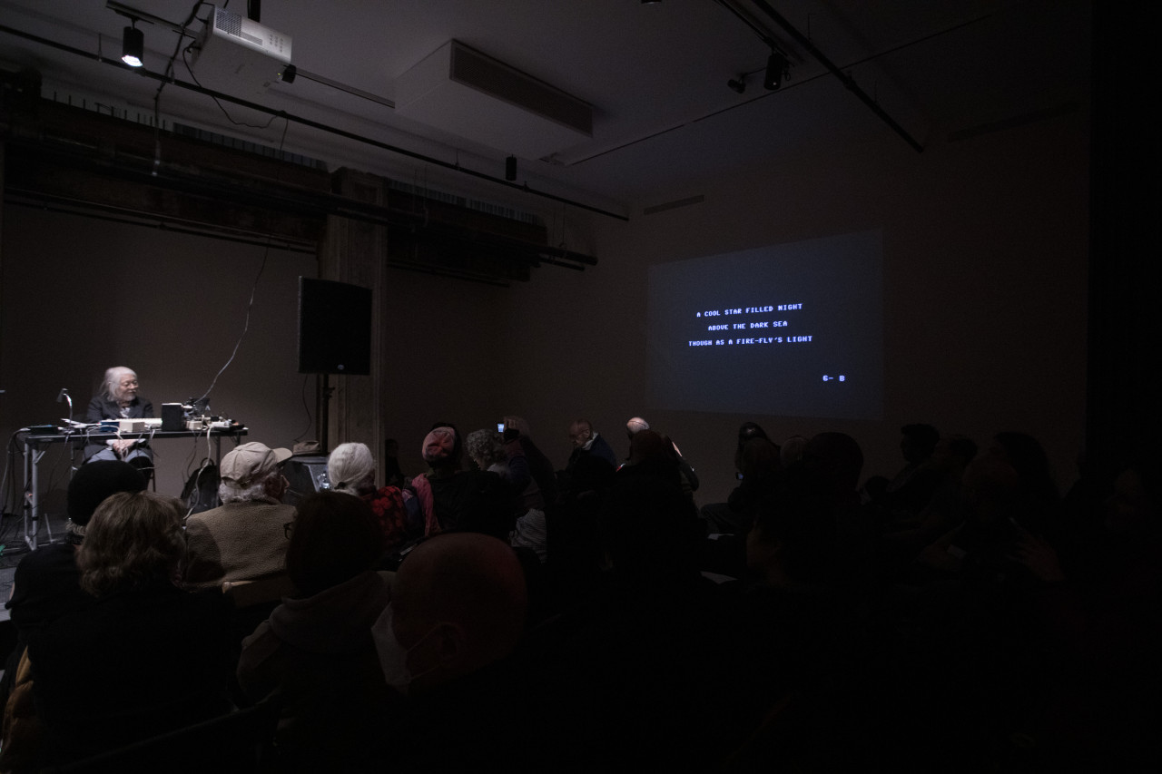 A crowd watches as a figure on stage sits behind a commodore computer while text is projected on the right wall that reads "A cool star filled night / above the dark sea / though as a fire-fly