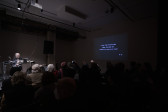 A crowd watches as a figure on stage sits behind a commodore computer while text is projected on the right wall that reads 